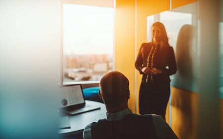 Silhouettes of two businesspeople during a meeting in a yellow board room: rearview of a man entrepreneur, his female colleague standing in a defocused background aloof a table with a laptop on itの写真素材
