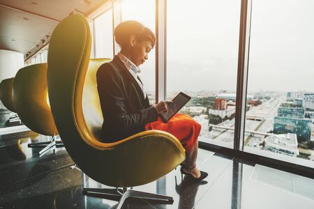 Side view of a dazzling young African-American woman entrepreneur sitting with a digital tablet on a yellow curved armchair indoors of a luxurious office room on a top floor of a business skyscraperの写真素材