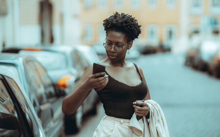 A fancy charming young African female in spectacles and a tank top is taken aback by an incoming message on her smartphone on the street; black girl with a cellphone outdoorsの写真素材