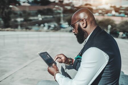An elegant mature bald African man with a beard and in spectacles is using outdoors his futuristic gadget with a user interface and infographics on it to implement a secure login using his fingerprintの写真素材