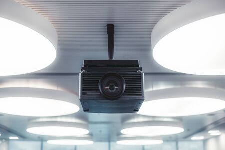 A black modern projector with a plastic case and a huge lens with aberrations in it mounted to a striped ceiling with multiple huge circle ceiling lamps lighting a modern conference hallの写真素材