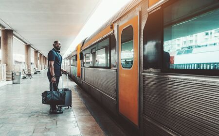 Wide-angle view of an elegant bearded bald mature African man entrepreneur standing with his bags on a railway station platform next to the door of a high-speed train, ready to start his business tripの写真素材