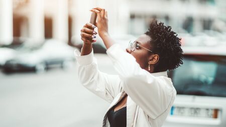 Side view of a young African female outdoors in a white cloak and spectacles holding a smartphone in her hands with nail-art and taking pics of something above using a rear camera of the gadgetの写真素材