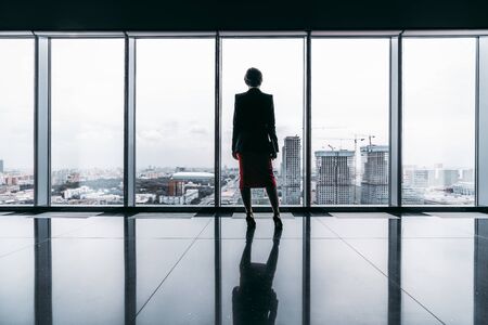 Rearview of a female entrepreneur on the top of a modern business office skyscraper standing near big panoramic windows and pensively looking at cityscape and construction site; floor with reflectionsの写真素材