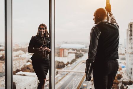 Two mature business partners near a panoramic window indoors of an office skyscraper: a man with a laptop and his caucasian female colleague with a smartphone; the cityscape and the highway outsideの写真素材