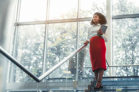 Dazzling African-American businesswoman with a tablet pc is standing on the top of a glass office staircase with chrome railing, a copy space area on the left for an ad text message or your logoの写真素材