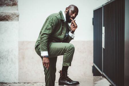 A fancy bald bearded black mature guy with a cigar and in a dark-green elegant suit and leather boots is resting his foot on a marble step in front of a wall and a fence and looking at the cameraの写真素材