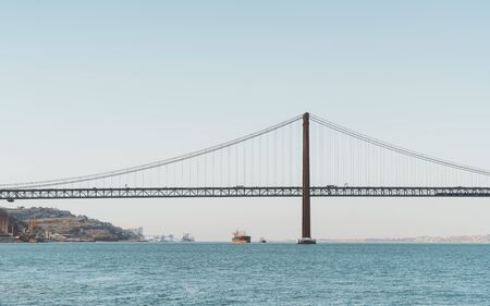 A view from the water of a huge suspension bridge "Ponte 25 de Abril" over the river Tagus in Lisbon, Portugal on a warm morning day with the horizon in the backgroundの写真素材