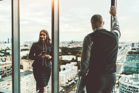 A meeting of two entrepreneurs on the top floor of a business skyscraper near the window: a businesswoman with a smartphone and her male partner back to the camera holding a laptop in his handの写真素材