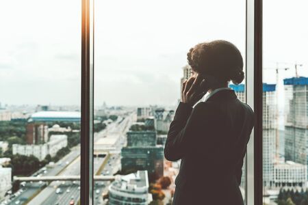 View from behind of a successful African-American woman entrepreneur talking on the cellphone near a panoramic window of a luxury office high-rise and looking at the cityscape with the highway belowの写真素材