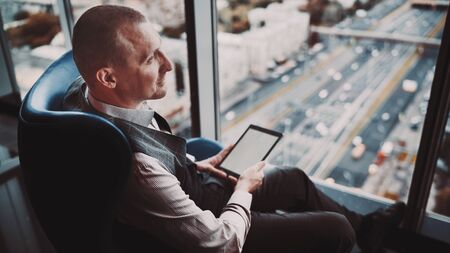 A handsome successful man entrepreneur with a tablet pc in hands is sitting on an armchair of a classic blue color on the top floor of a luxurious business high-rise with a high-way outside the windowの写真素材