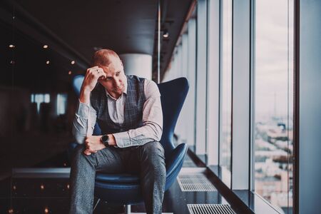 An annoyed and depressed caucasian man entrepreneur is sitting on an armchair of classic clue color in his business office skyscraper and sadly looking outside the window on the cityscape belowの写真素材