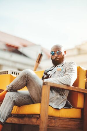 A vertical shot an elegant mature bearded bald African man in round bluish sunglasses and a...
