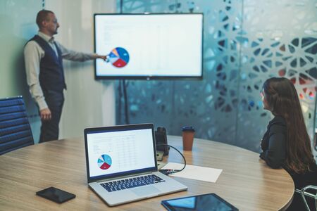 A business meeting in a board room with selective focus on a laptop and gadgets in the foreground and two partners in the background: man entrepreneur is showing on the diagram on the screen of plasmaの写真素材