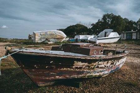 An abandoned wooden fishing boat with a small cabin and flacked timbers on a pebble ground of a shallowed pier on an overcast day surrounded by other boats in the distance, Alcochete, Portugalの写真素材
