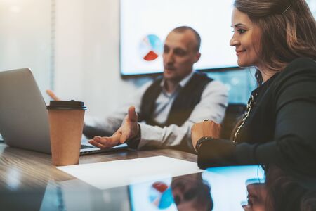 Business meeting in an office board room: a charming adult woman entrepreneur is listening to a financial report of her partner in the defocused background, laptop and paper cup on the table in frontの写真素材