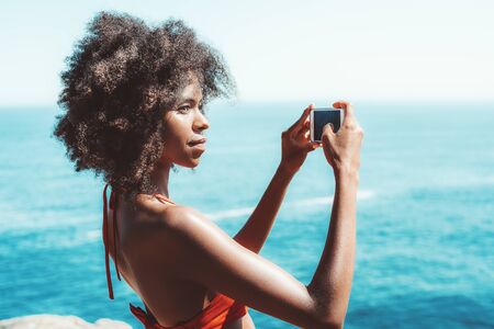 Portrait of a dazzling African-American girl shooting a seascape on her smartphone; young charming Brazilian female with a curly Afro hair and in swimwear is photographing resort using the cellphoneの写真素材
