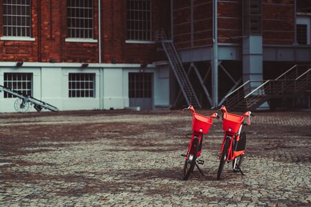 Two vivid red rental bikes parked on a paving stone in front of a brick building; two shared bicycles with metal baskets at the helm standing outdoors on the pavement, with copy space area on the leftの写真素材