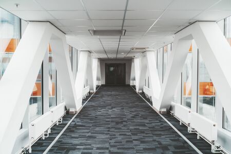 Bright long modern passageway between two towers of an office or a hospital skyscraper with  zig-zag lines of the load-bearing beams, suspended ceiling, heating batteries, and carpeting on the floorの写真素材