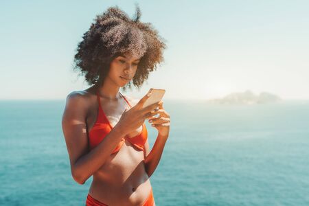 Dazzling young African-American female with curly afro hair standing in a swimsuit on a beach with ocean and a small island in the background and using her smartphone; a copy space place on the rightの写真素材