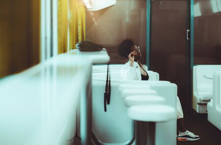 An elegant charming young African female in spectacles in a dining car of a modern high-speed train; a fancy black woman in a white trench adjusting her eyeglasses while sitting in a restaurant carの写真素材