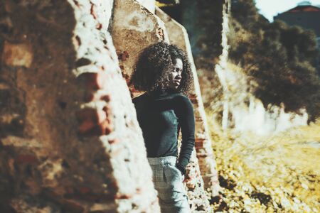 Dazzling black girl with curly hair standing in between ofÂ old brick ledges and looking aside;...