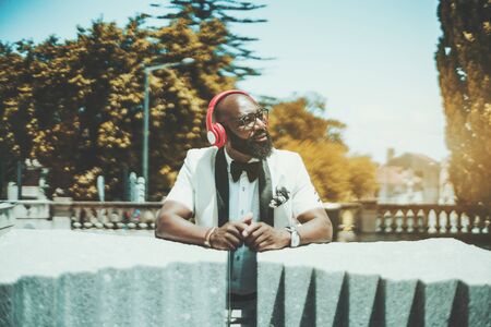 Handsome mature bearded bald African man in glasses and an elegant white suit is enjoying sunny...