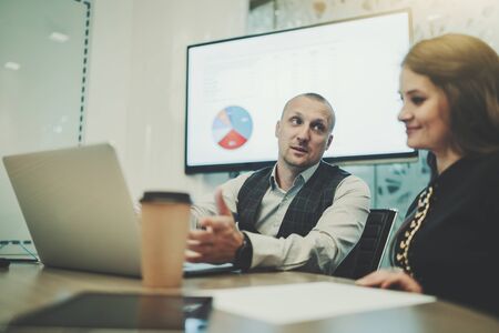 Business meeting in a boardroom with a selective focus on a caucasian man entrepreneur talking with his female colleague; a laptop, paper coffee cup, and a paper sheets on the table, LCD screen behindの写真素材