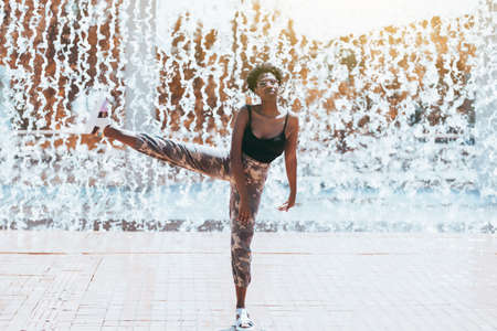 Funny shot with a fancy charming black girl in a sunglasses fooling around, standing on one leg with another leg lifted up doing ballet pas; streams of falling water in background and splashes aroundの写真素材