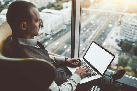 Side view of a stately successful man entrepreneur with a laptop with white blank screen mock-up,...