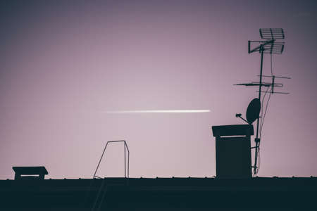 A dark late evening shot with selective focus on a silhouette of the roof with antennas and ventilation pipes above in the foreground and a white trace of an airplane in a purple defocused sky behindの写真素材