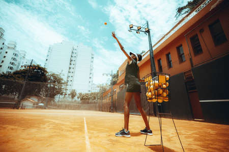 Wide-angle side view of a svelte African-American girl having a tennis training on a bright outdoor court, with a basket of orange balls near her and a copy space place on the left for an ad messageの写真素材