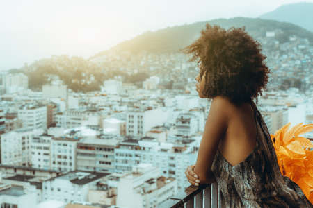 Side view of thoughtful sleepy an African-American girl leaning against a balcony fence and pensively observing typical hilly Brazilian cityscape on a rainy morning sunrise during a quarantineの写真素材
