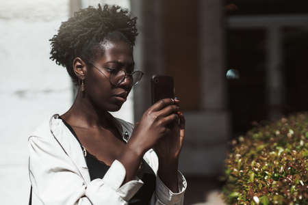 Portrait of a young beautiful black woman in a white trench and spectacles taking pics outdoors...
