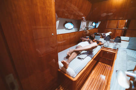 A rich carefree bearded black guy in shorts is laying on a bed in a cabin of a luxury yacht with a...