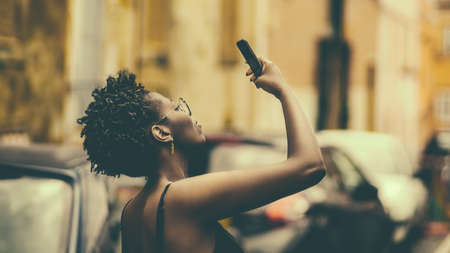 Side view of a young black female using her smartphone to take photos on the street, retro colors;...