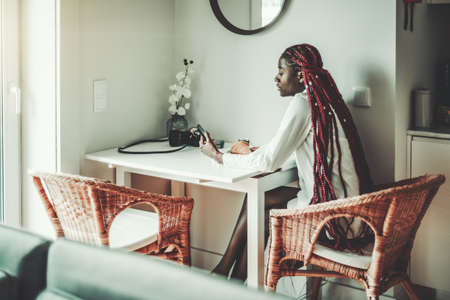 A ravishing pensive young black female with long braidsÂ of a dark-red color is sitting on a wicker wooden armchair at the kitchen table in the morning during breakfast and using her smartphoneの写真素材