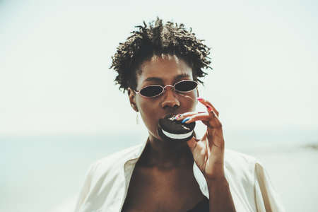 Portrait of a young beautiful black woman in sunglasses and a white trench, holding in her hand with...