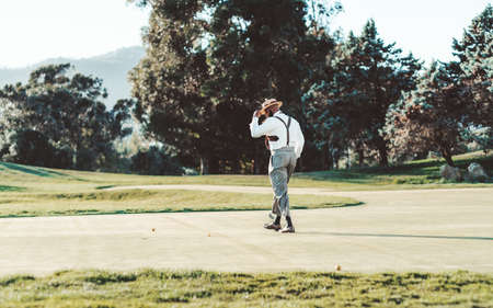 View of a golf field on a sunny windy day with an elegant retrosexual black guy in a hat, white shirt, and suspender trousers, he is walking to a ball on the grass to hit it towards the closest holeの写真素材