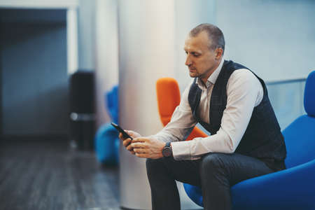 A handsome mature caucasian man entrepreneur is sitting on a blue armchair indoors of an office hall and reading messages on the screen of his phone, with a copy space place on the left for an ad textの写真素材