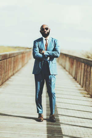 Vertical shot of a handsome mature bald black man entrepreneur in a formal elegant custom made blue suit, sunglasses, necktie; he is standing in the middle of a long wooden bridge with hands crossedの写真素材