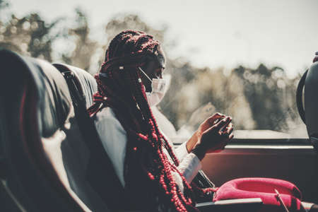 A black woman in a virus protective face mask and with long braided hair of red color is sitting on a leather seat of a regular intercity bus and watching an online show using her smartphoneの写真素材