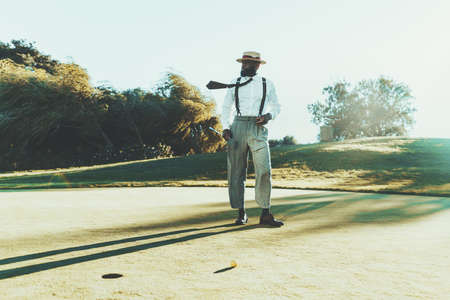 A fashionable mature bearded black guy in a hat, white shirt, and trousers with suspenders is holding in his hands a cigar and a golf club while standing on a golf field next to the ball and the holeの写真素材