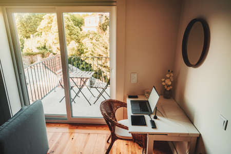 A cozy beautiful nook lit with warm light with a desktop, there are laptop, a USB hub, a smartphone, and a wireless graphic tablet on the table, a balcony with a view on a sunny day in the backgroundの写真素材