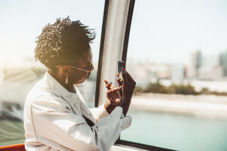 Side view of charming young African woman tourist with curly black hair inside of a ropeway cabin, she is holding a smartphone in her hands with nail-art and taking pictures of sights and selfies tooの写真素材