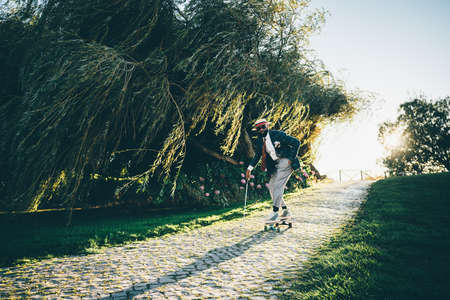 A fancy handsome black man in a hat, elegant suit with trousers and necktie, with a golf club in his hand, is riding a skateboard and descending from a small hill paved with stones on a sunny eveningの写真素材