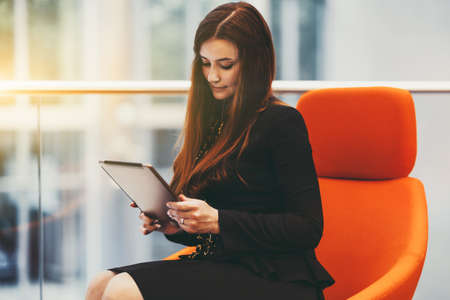Portrait of a charming mature caucasian woman entrepreneur in a black strict costume having a group video call using her digital tablet while sitting in an office open-space area on an orange armchairの写真素材