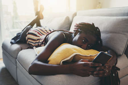 A young charming relaxed black female with braided hair is laying in the living room of her apartment on a sofa and yellow cushion and chatting with friends using her smartphone, a sunny morningの写真素材