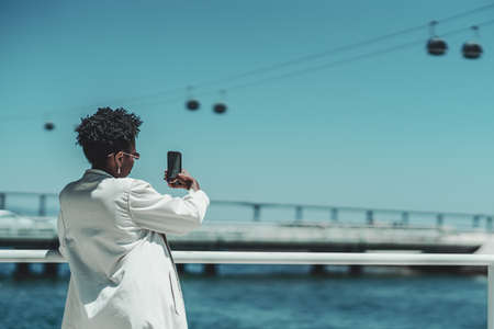 View from behind of an elegant young black female with curly afro hair in stylish sunglasses and a white trench is standing outdoors and taking a selfie or photographing on the cellphone a ropewayの写真素材