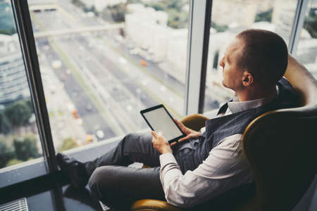 Side view of a pensive mature man entrepreneur in a plaid vest, sitting near the window on an orange armchair in an office cabinet on a top floor of a business high-rise with a digital tablet in handsの写真素材